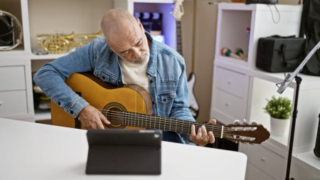 Un homme mûr joue de la guitare en regardant un tutoriel sur une tablette dans un bureau à domicile bien éclairé.