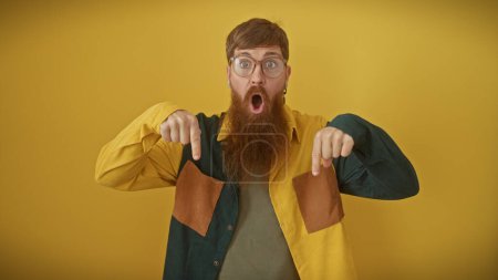 Shocked man with beard pointing down while standing against a bright yellow background