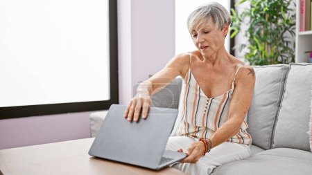 Téléchargez les photos : Une femme mûre aux cheveux gris courts s'assoit sur un canapé tout en fermant son ordinateur portable dans un salon confortable, symbolisant l'équilibre travail-vie privée. - en image libre de droit