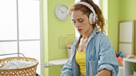 Foto de Una mujer joven escucha música en los auriculares mientras hace la colada en un interior verde y luminoso. - Imagen libre de derechos