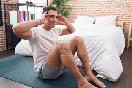 Téléchargez les photos : Jeune homme hispanique souriant entraînement confiant abs exercice dans la chambre - en image libre de droit