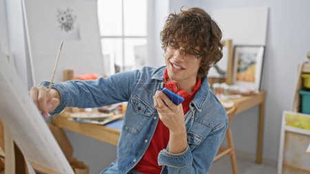 Téléchargez les photos : Un jeune artiste dans un studio intérieur multitâches avec un pinceau et un smartphone, exsudant créativité et concentration. - en image libre de droit