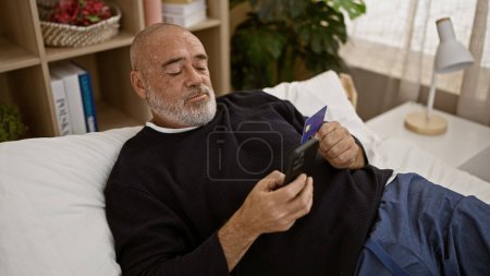 A mature bearded man examines documents in a well-lit, cozy bedroom, representing a moment of thought or financial planning.