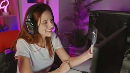 Eine fröhliche junge Frau mit roten Haaren macht ein Selfie in einem neonbeleuchteten Spielzimmer zu Hause, trägt Kopfhörer.