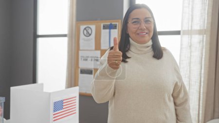 Mujer hispana sonriente dando pulgares arriba en un centro de votación de EE.UU. con cabina de votación y bandera americana.