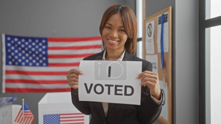 Foto de Mujer afroamericana sonriente sosteniendo el letrero 'voté' en un centro electoral con nuestra bandera - Imagen libre de derechos