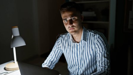 Téléchargez les photos : Un beau jeune homme dans son bureau éclairé par une lampe de bureau la nuit, se concentrant sur son ordinateur portable. - en image libre de droit