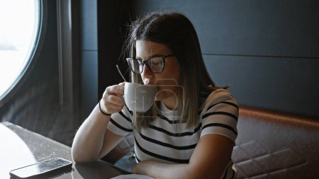 Téléchargez les photos : Jeune femme hispanique dégustant un café à une table de café avec smartphone, lunettes et vue sur la mer. - en image libre de droit