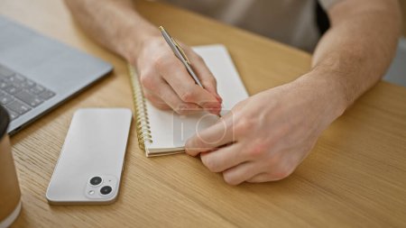 Photo for A man writes in a notebook next to his smartphone and laptop in a modern office setting. - Royalty Free Image