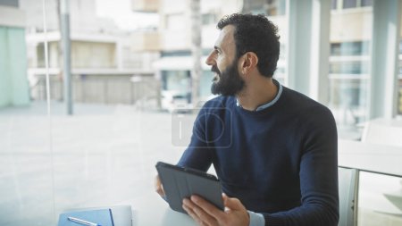 Photo for Bearded man looks pensively out of a window in an office, holding a tablet, with notebook and urban background visible. - Royalty Free Image