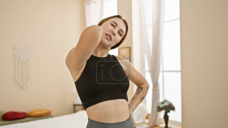Foto de Una joven morena hace muecas de dolor mientras sostiene su cuello, de pie en un dormitorio naturalmente iluminado con decoración en el fondo. - Imagen libre de derechos