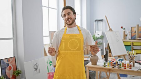 Téléchargez les photos : Jeune homme hispanique artiste tenant pinceau et palette souriant au studio d'art - en image libre de droit