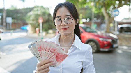 Téléchargez les photos : Une jeune femme se tient dans une rue de la ville, affichant en toute confiance un fan de billets de banque en yuan chinois. - en image libre de droit
