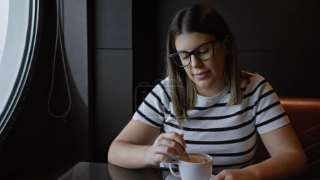 Téléchargez les photos : Femme hispanique adulte dans des lunettes brassant du café à une table de café près d'une fenêtre, évoquant calme et loisirs. - en image libre de droit