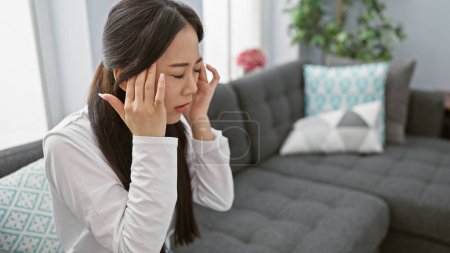Foto de Una mujer china estresada con dolor de cabeza en casa, sentada en un sofá en una sala de estar moderna. - Imagen libre de derechos
