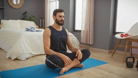 Photo for A bearded man in sportswear practicing yoga on a mat in a modern bedroom, symbolizing wellness and self-care. - Royalty Free Image