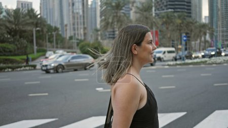 Photo for A young brunette woman wearing casual clothing stands by a crosswalk amidst the modern skyscrapers of dubai's cityscape. - Royalty Free Image