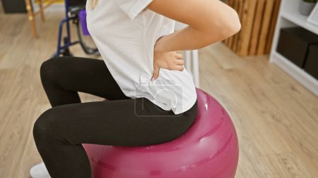 Young woman experiencing back pain while sitting on a fitness ball indoors at a clinic.