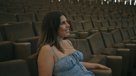 A young, smiling hispanic woman sits alone in a spacious theater, exuding a sense of leisure and entertainment.