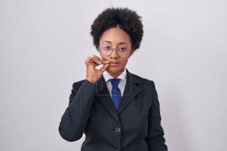 Beautiful african woman with curly hair wearing business jacket and glasses mouth and lips shut as zip with fingers. secret and silent, taboo talking 