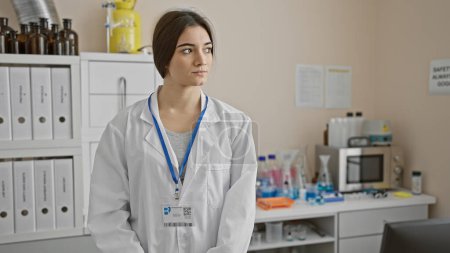 Foto de Una joven técnica hispana con bata de laboratorio se encuentra pensativamente en un ambiente de laboratorio, insinuando profesionalismo médico. - Imagen libre de derechos