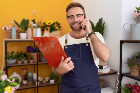 Téléchargez les photos : Middle age man florist talking on smartphone reading clipboard at flower shop - en image libre de droit