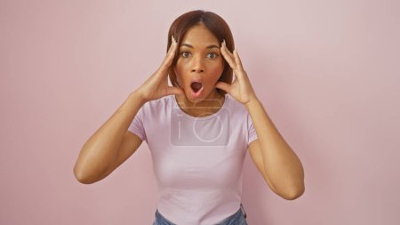 Téléchargez les photos : Une femme afro-américaine surprise en tenue décontractée sur fond rose exprimant son étonnement avec les mains sur les joues. - en image libre de droit