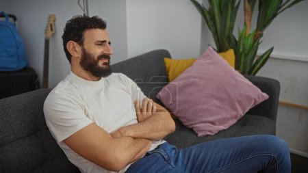 Photo for Middle-aged bearded man with crossed arms sits thoughtfully on a couch in a modern living room. - Royalty Free Image