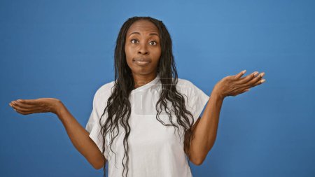 Photo for A puzzled young african american woman with curly hair wearing a white shirt against a blue background shrugs with an uncertain expression. - Royalty Free Image