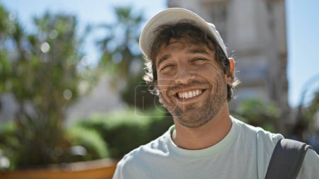 Téléchargez les photos : Bel homme hispanique souriant avec barbe et les yeux verts portant un chapeau à l'extérieur en milieu urbain - en image libre de droit