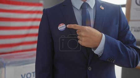 Photo for A man in a suit points to a 'voted' sticker with an american flag backdrop in a voting center. - Royalty Free Image