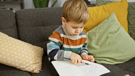 Photo for A focused blonde toddler boy is doodling in a notebook while sitting on a couch inside a cozy living room. - Royalty Free Image