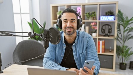 Hombre hispano guapo con una barba sonriendo mientras organiza un espectáculo en el interior de un estudio de radio.