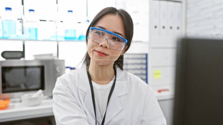 A young asian woman wearing a lab coat and safety glasses stands in a laboratory setting.