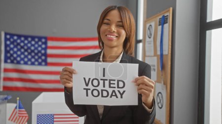 Smiling african american woman proudly holds an 'i voted today' sign in a us polling center with a flag background.