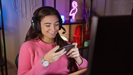 Téléchargez les photos : Une femme souriante portant un casque utilise un smartphone dans une salle de jeux éclairée au néon la nuit - en image libre de droit