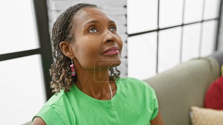 Foto de Una mujer afroamericana reflexiva en su sala de estar, mirando hacia arriba con esperanza y aspiración - Imagen libre de derechos