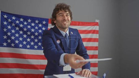 Téléchargez les photos : Un jeune hispanique barbu, vêtu d'un costume, prolonge un bulletin de vote dans un collège électoral américain orné d'un drapeau. - en image libre de droit