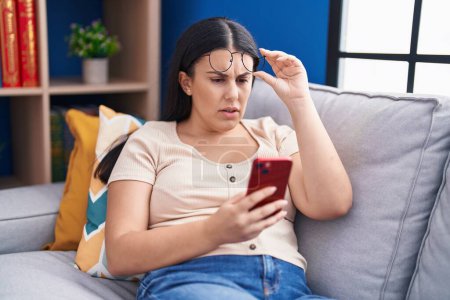 Foto de Joven hermosa mujer hispana mirando teléfono inteligente de la pantalla con problema de visión en casa - Imagen libre de derechos