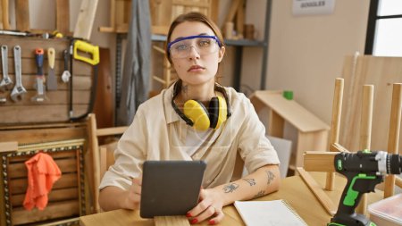 Photo for A focused brunette woman wearing safety glasses and headphones holds a tablet in a carpentry workshop. - Royalty Free Image