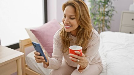 Photo for Smiling hispanic woman enjoying morning coffee while browsing smartphone in a cozy bedroom setting - Royalty Free Image