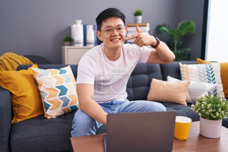 Téléchargez les photos : Jeune homme asiatique à l'aide d'un ordinateur portable à la maison assis sur le canapé souriant gai montrant et pointant avec les doigts les dents et la bouche. concept de santé dentaire. - en image libre de droit