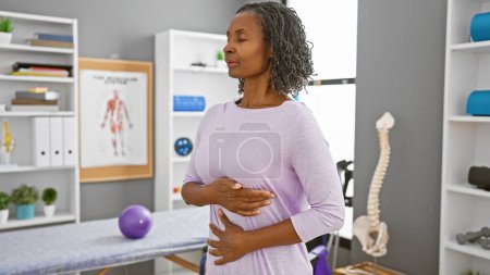 Une femme noire d'âge moyen pratique des exercices respiratoires dans une clinique de réadaptation, entourée d'équipement thérapeutique.