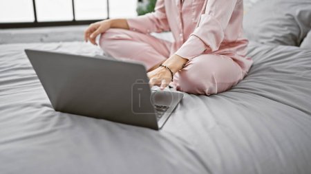 Foto de Mujer joven en pijama usando portátil mientras está sentada con las piernas cruzadas en la cama en un dormitorio moderno - Imagen libre de derechos