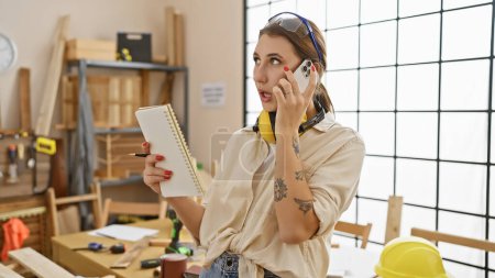 Photo for A young woman multitasks in a carpentry workshop, holding a notebook and talking on the phone. - Royalty Free Image