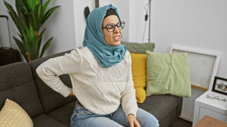 Photo for Mature muslim woman in hijab experiencing back pain while sitting on a couch indoors. - Royalty Free Image