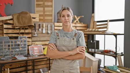 Photo for Confident young woman in a carpentry workshop, wearing an apron and standing with arms crossed. - Royalty Free Image
