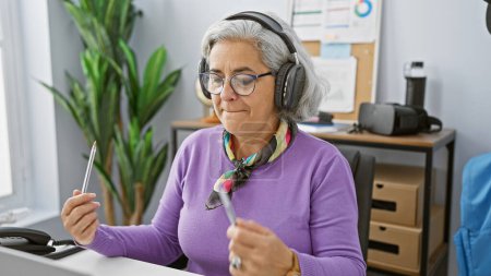 Foto de Una mujer de pelo gris con auriculares disfruta de la música mientras sostiene un bolígrafo en un entorno de oficina moderno. - Imagen libre de derechos