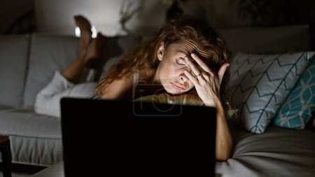 Tired woman on couch at night with laptop stresses in cozy home.
