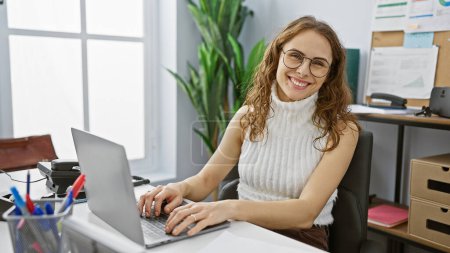 Photo for Smiling young hispanic woman at her desk in a bright modern office interior. - Royalty Free Image